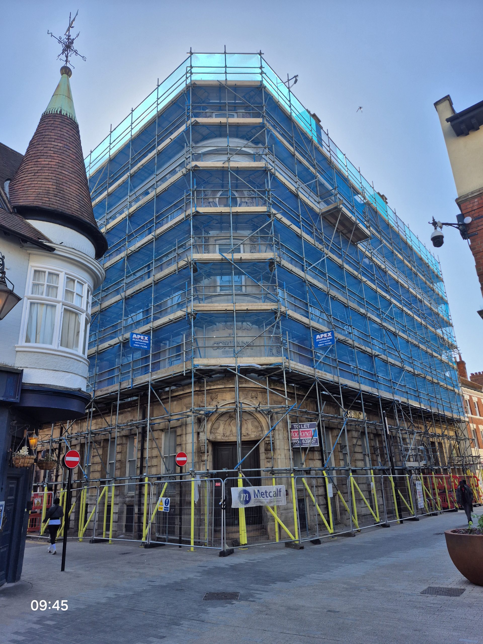 Apex Scaffolding installation on a multi-story historic corner building in Northampton town centre, featuring full blue debris netting, secure pedestrian hoarding, and yellow protective upright sleeves for a high-street commercial renovation project.